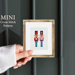 Może przedstawiać: Wzorzec haftu krzyżykowego w ramie przedstawiający dwóch żołnierzyków. Żołnierz po lewej stronie ma na sobie czerwony mundur ze złotymi ozdobami i czarny kapelusz. Żołnierz po prawej stronie ma na sobie różowy mundur z czarnym kapeluszem.