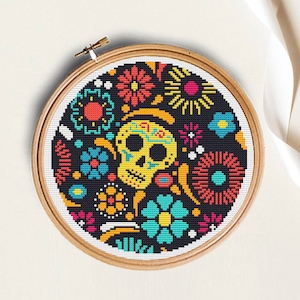 K&ouml;nnte beinhalten: Ein schwarzer und wei&szlig;er Kreuzstich-Stickrahmen mit einem farbenfrohen Zucker Sch&auml;del Design. Der Sch&auml;del ist von Blumen und anderen dekorativen Elementen umgeben.