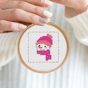 Puede incluir: Un aro de bordado de punto de cruz blanco con un diseño de muñeco de nieve rosa. El muñeco de nieve lleva un gorro y una bufanda rosas.
