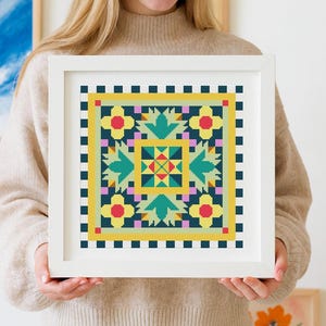 Op de afbeelding: Een ingelijst kruissteekkunstwerk met een geometrisch ontwerp in geel, turquoise, roze en marineblauw. Het vierkante ontwerp wordt door een persoon vastgehouden. Het kunstwerk bevat bloem- en geometrische elementen.