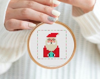 May include: A round, wooden-framed embroidery hoop with a white fabric background. The fabric features a cross-stitch design of Santa Claus wearing a red suit, white beard, and a red hat.