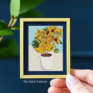 Peut inclure: Un motif de point de croix encadré représentant un vase de tournesols. Les tournesols sont jaunes et bruns, et le vase est blanc. Le cadre est doré.