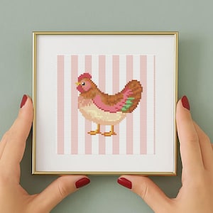 Könnte beinhalten: Ein gerahmtes Kreuzstich-Kunstwerk mit einem Cartoon-Huhn. Das Huhn ist braun, rosa und gelb, vor einem rosa-weiß gestreiften Hintergrund. Der Rahmen ist goldfarben, und das Kunstwerk wird von Händen mit rotem Nagellack gehalten.