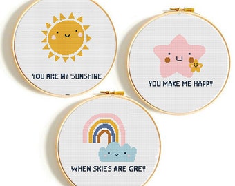 May include: Three embroidery hoop art pieces with cute illustrations and text. The first hoop features a yellow sun with a smiling face and the text "You are my sunshine". The second hoop features a pink star with a smiling face and the text "You make me happy". The third hoop features a rainbow and a blue cloud with a smiling face and the text "When skies are grey".