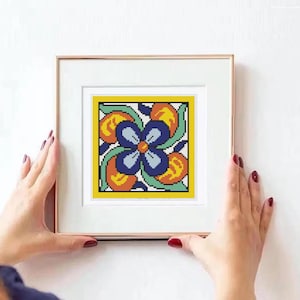 May include: A framed cross-stitch artwork featuring a symmetrical floral design. The design includes a central blue flower, surrounded by orange and yellow accents, all within a yellow border. The frame is gold-colored.