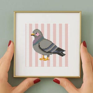 May include: A square gold-toned frame containing a cross-stitch embroidery of a grey pigeon with pink cheeks and yellow feet, set against a background of vertical white and light pink stripes. The artwork is centered within the white matting.