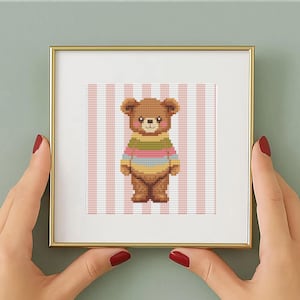 May include: A framed cross-stitch artwork featuring a brown teddy bear wearing a striped sweater in shades of green, pink, and blue. The background has pink and white vertical stripes. The frame is gold-colored.
