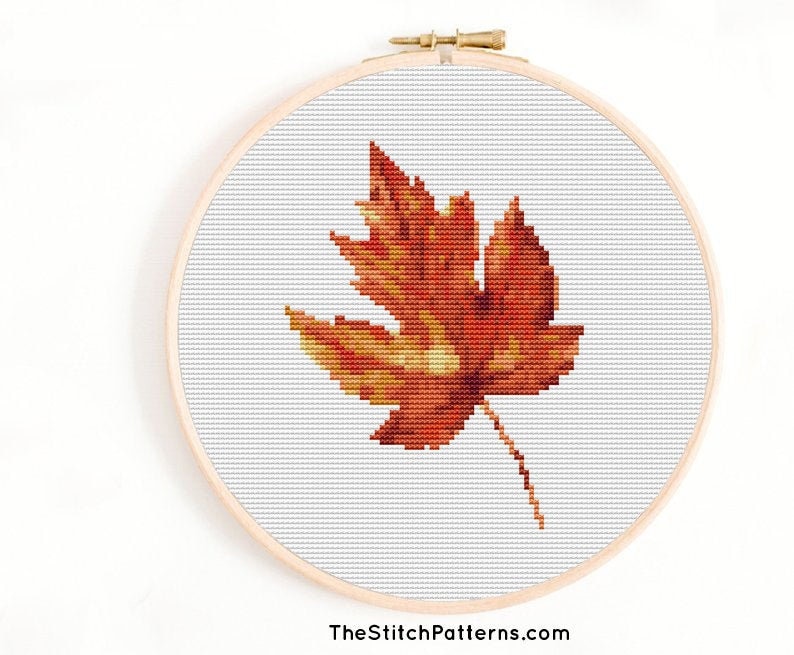 May include: A cross-stitch pattern of a single orange and yellow autumn leaf on a white background.