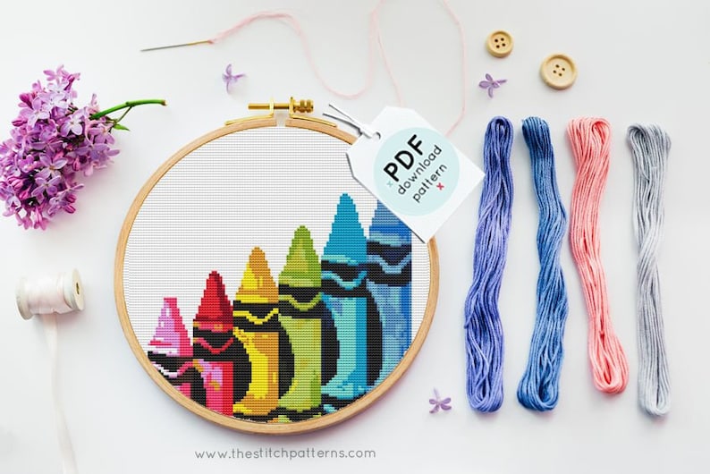 May include: A cross-stitch embroidery hoop with a design of colorful crayons. The hoop is on a white surface with embroidery floss and buttons.