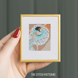 May include: A framed cross-stitch pattern of a dancer in a blue and white dress with orange and brown accents. The dancer is in mid-air with her arms outstretched.