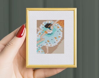 May include: A framed cross-stitch pattern of a dancer in a blue and white dress with orange and brown accents. The dancer is in mid-air with her arms outstretched.