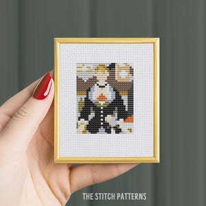 May include: A framed cross-stitch pattern of a person in a black jacket with a red flower on their chest. The background is a blurry image of a building.