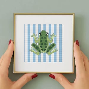 May include: A framed cross-stitch art piece featuring a green frog with a white and blue striped background. The frog is detailed with various shades of green. The frame is gold-colored.