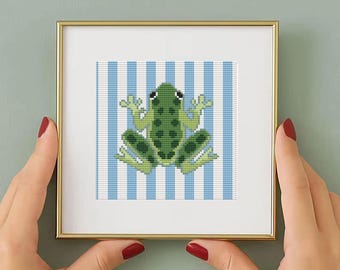 Puede incluir: Una obra de arte en punto de cruz enmarcada que presenta una rana verde sobre un fondo de rayas azules y blancas. La rana está detallada con varios tonos de verde. El marco es dorado.