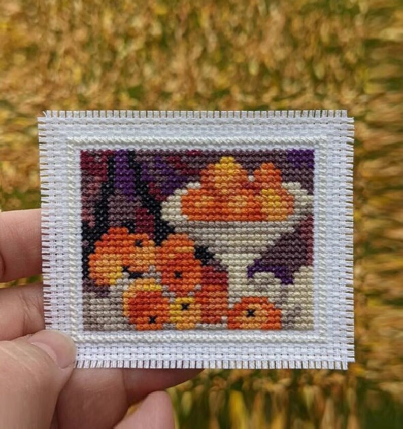 May include: A cross-stitch embroidery of a bowl of orange pumpkins with a purple and white background.