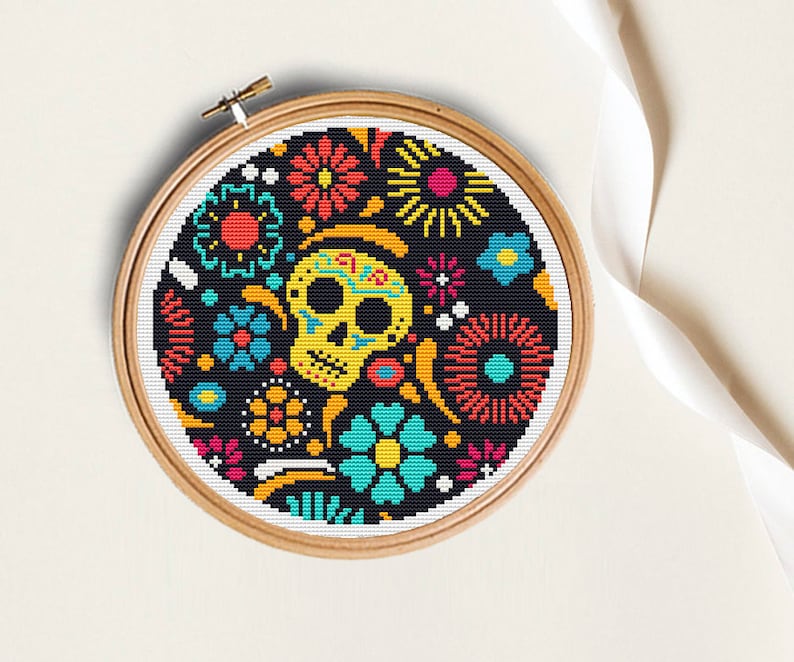 K&ouml;nnte beinhalten: Ein schwarzer und wei&szlig;er Kreuzstich-Stickrahmen mit einem farbenfrohen Zucker Sch&auml;del Design. Der Sch&auml;del ist von Blumen und anderen dekorativen Elementen umgeben.
