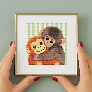 May include: A framed cross-stitch artwork featuring two hugging monkeys. One monkey is orange, the other is brown. The background has green and white vertical stripes. The frame is gold.