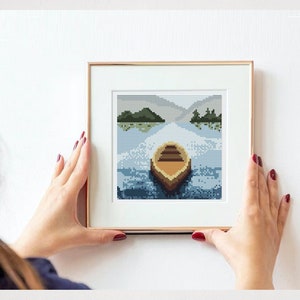 Puede incluir: Una obra de arte de punto de cruz enmarcada que representa un pequeño bote de madera en un lago con islas verdes en el fondo.