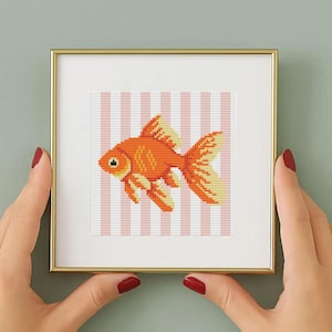 May include: A square gold-toned frame containing a cross-stitch embroidery of an orange goldfish. The fish is depicted in profile against a background of vertical white and light pink stripes. The embroidery features detailed stitching.
