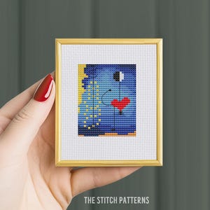 Op de afbeelding: Een ingelijst kruissteekpatroon met een abstract ontwerp in blauw, geel, rood, zwart en wit. Het ontwerp bevat een hart, een cirkel en een lijn.