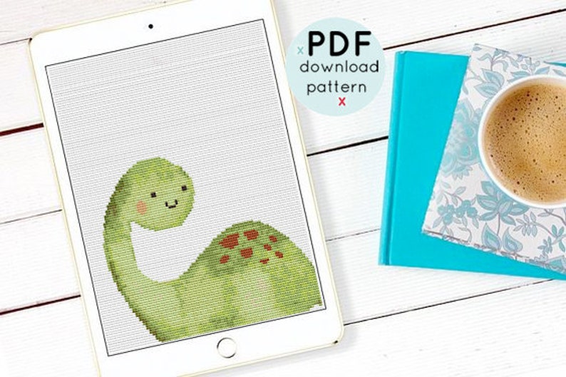 May include: A digital tablet displaying a cross-stitch pattern of a green dinosaur with red spots. The text "PDF download pattern" is displayed on the screen.