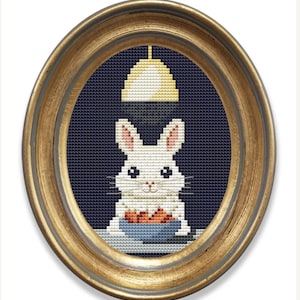 May include: A white rabbit sits under a yellow light fixture and eats from a blue bowl filled with orange carrots. The rabbit is framed in a gold oval.