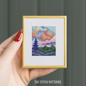 Pode incluir: Um padrão de ponto cruz de uma paisagem com uma árvore azul, nuvens cor-de-rosa e colinas verdes. O padrão é emoldurado em uma moldura dourada.