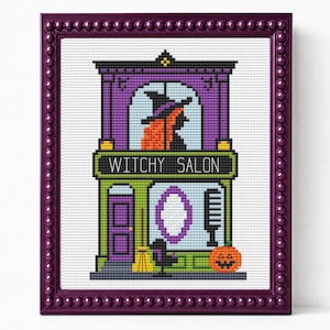 May include: A cross-stitch embroidery in a dark purple beaded frame depicting a two-story building labeled WITCHY SALON. The design features a witch in the upper window, a purple door, a mirror, a comb, and a jack-o'-lantern on the ground floor.
