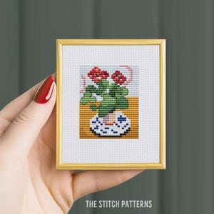 May include: A framed cross-stitch artwork featuring a potted plant with red flowers and green leaves. The plant sits in a blue and white patterned pot against a yellow and white background. The frame is gold. Text at the bottom reads "THE STITCH PATTERNS."