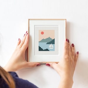 May include: A framed cross-stitch artwork depicting a stylized landscape with a red sun, blue water, and green mountains. The frame is a light brown color.