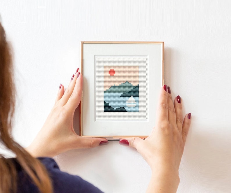 May include: A framed cross-stitch artwork depicting a stylized landscape with a red sun, blue water, and green mountains. The frame is a light brown color.