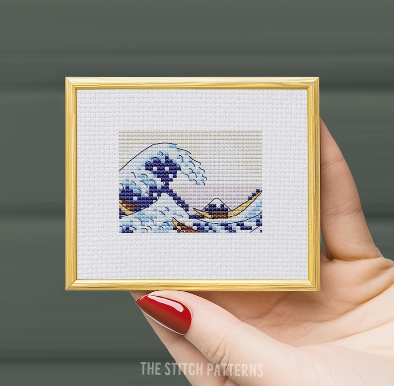 Puede incluir: Un patr&oacute;n de punto de cruz enmarcado de la Gran Ola de Kanagawa, una famosa estampa japonesa en madera de Hokusai. La ola est&aacute; representada en tonos de azul, blanco y amarillo, con una monta&ntilde;a en el fondo.