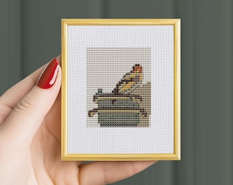 May include: A cross-stitch pattern featuring a yellow bird with a red head perched on a wooden structure. The pattern is framed in a gold frame.