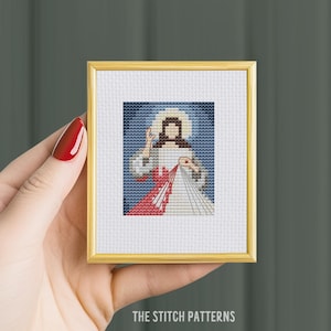 Puede incluir: Un patrón de punto de cruz enmarcado de Jesucristo con un corazón rojo y rayos de luz. El patrón está en un marco dorado y se sostiene en una mano.