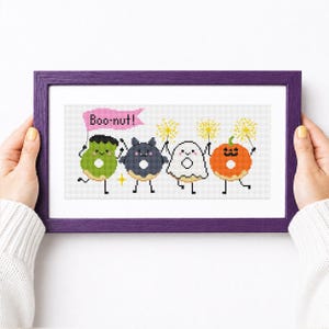 May include: A cross-stitch pattern in a purple frame featuring four anthropomorphic donuts decorated as a green monster, a dark gray bat, a white ghost, and an orange pumpkin. The design includes yellow sparklers and a pink flag with the text Boo-nut!