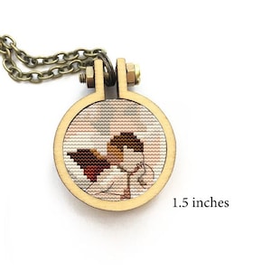 May include: A wooden embroidery hoop necklace featuring a cross-stitch design of a person with wings. The hoop is 1.5 inches in diameter.
