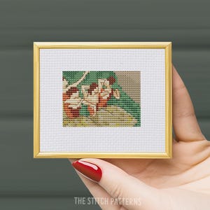 May include: A framed cross-stitch pattern depicting three figures in a swimming pool. The figures are wearing red and white bathing suits and are surrounded by green and blue water. The cross-stitch is in a gold frame.