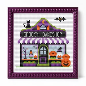 May include: A cross-stitch embroidery of a bakeshop in a purple beaded frame. The design features a purple and black building labeled SPOOKY BAKESHOP, with a black cat, a bat, pumpkins, and a striped awning on a white fabric background.