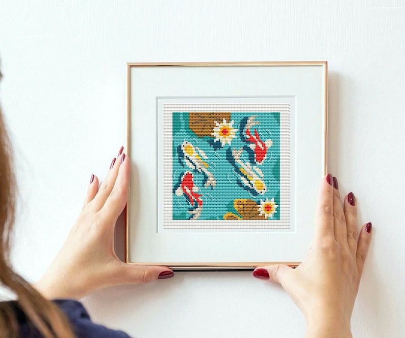 May include: A framed cross-stitch pattern featuring a blue pond with white and red koi fish swimming amongst lily pads and flowers.