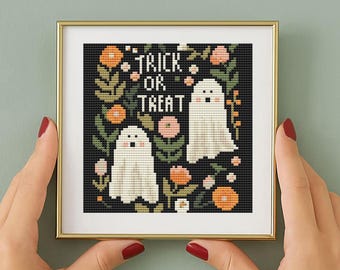 Puede incluir: Un bordado de punto de cruz enmarcado con dos fantasmas amigables y las palabras "Trick or Treat" sobre un fondo negro. El diseño incluye flores naranjas y rosas y follaje verde, todo dentro de un marco dorado. La obra de arte está sostenida por manos con esmalte de uñas rojo.
