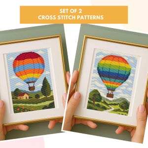 May include: Two framed cross-stitch patterns depicting hot air balloons in vibrant colours. The balloons are striped with red, orange, yellow, green, and blue. The frames are gold-coloured with white mounts. The background shows a landscape with trees and a house. Text reads: SET OF 2 CROSS STITCH PATTERNS.