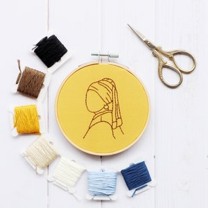 Pode incluir: Um aro de bordado amarelo com um desenho de linha marrom de uma mulher com um brinco de p&eacute;rola. O aro &eacute; cercado por carret&eacute;is de fio de v&aacute;rias cores, incluindo marrom, amarelo, branco, azul claro e azul escuro.