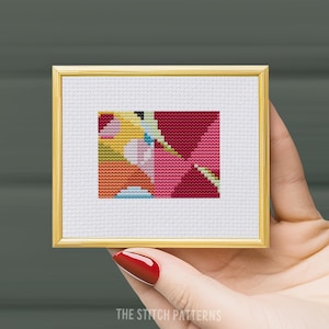 May include: A framed cross-stitch pattern with a geometric design featuring pink, red, yellow, orange, and blue colors.