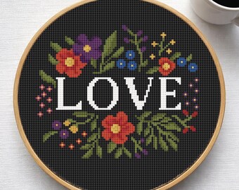 Peut inclure: Un cercle de broderie au point de croix avec le mot "LOVE" en blanc, entouré d'une couronne florale. Les fleurs sont rouges, violettes et bleues, avec des feuilles vertes, sur fond noir. Le cercle en bois est visible.