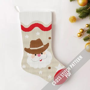 May include: A cross stitch pattern for a Christmas stocking featuring a Santa Claus wearing a cowboy hat. The stocking is beige with red and white accents. The pattern is available as a PDF download.