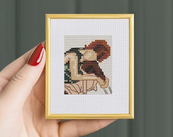 May include: A framed cross-stitch pattern of a woman in a green dress with red hair, sitting on a chair. The cross-stitch is in a gold frame.