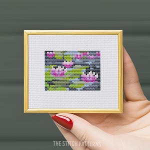 Peut inclure: Un motif de point de croix encadré représentant des nénuphars roses dans un étang. Le motif est brodé sur une toile Aida blanche et encadré dans un cadre doré.