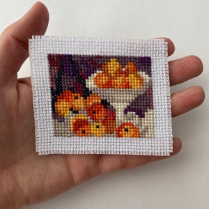 May include: A cross-stitch embroidery of a bowl of oranges, with a white background and a purple and brown border.