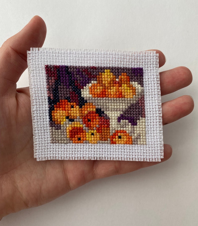 May include: A cross-stitch embroidery of a bowl of oranges, with a white background and a purple and brown border.