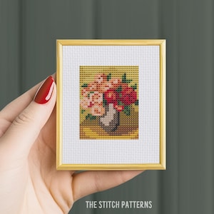 以下が含まれることがあります： 白い花瓶にピンクと赤の花束が描かれた、額装されたクロスステッチ刺繍。花は黄色い背景の花瓶にアレンジされています。